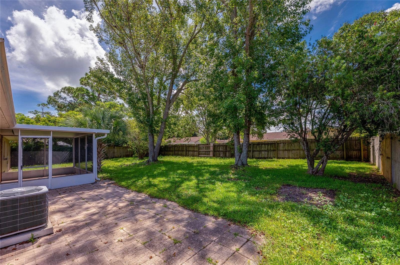 156 SPRINGWOOD DRIVE, Daytona Beach, FL 32119 Spacious backyard featuring an open paved patio, a fully enclosed screen porch, and an exterior air conditioning unit. Mature trees and a wooden perimeter fence enclose the lush green lawn.