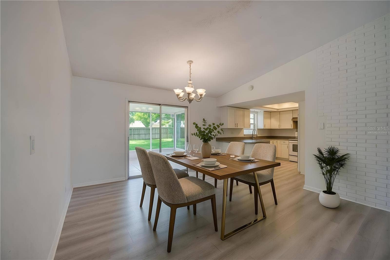 156 SPRINGWOOD DRIVE, Daytona Beach, FL 32119 Virtually Staged. Open dining and living space defined by a classic chandelier, light luxury vinyl flooring, and a white brick wall accent. Sliding glass doors open to a screened porch, filling the space with natural light.