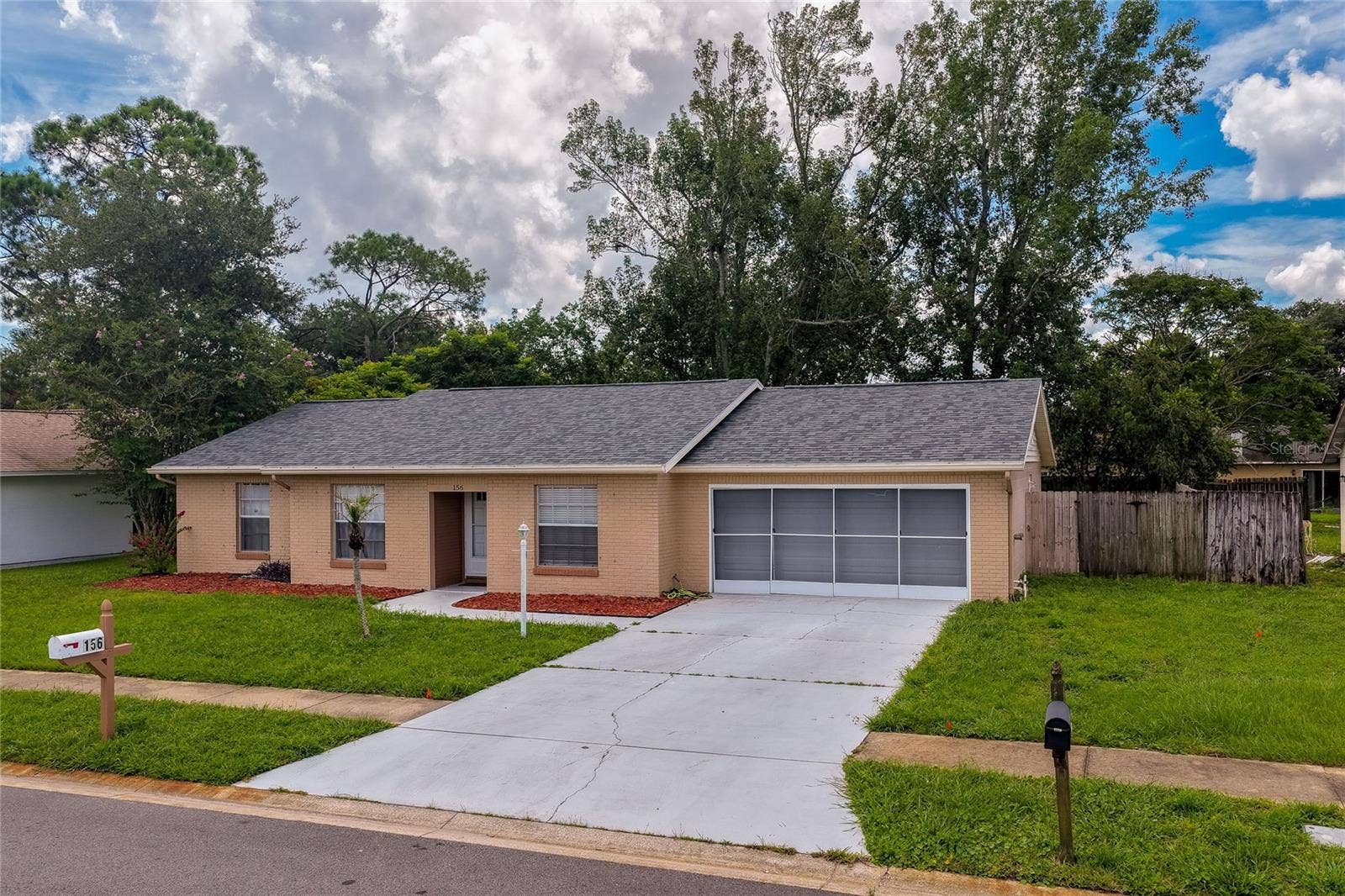 156 SPRINGWOOD DRIVE, Daytona Beach, FL 32119 Front-facing exterior view highlighting a wide concrete driveway, a screened multi-car garage, and a neatly manicured front yard. A wood post mailbox sits near the street curb alongside lush green lawn landscaping.