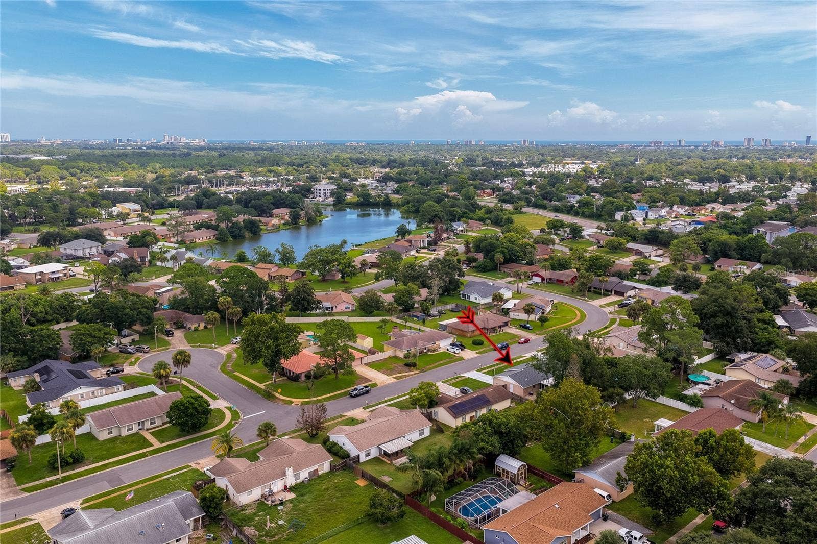 156 SPRINGWOOD DRIVE, Daytona Beach, FL 32119 Broad aerial photo showing the home situated within a quiet suburban development near a scenic local lake. Distant views capture the sprawling green canopy extending toward the coastal horizon.