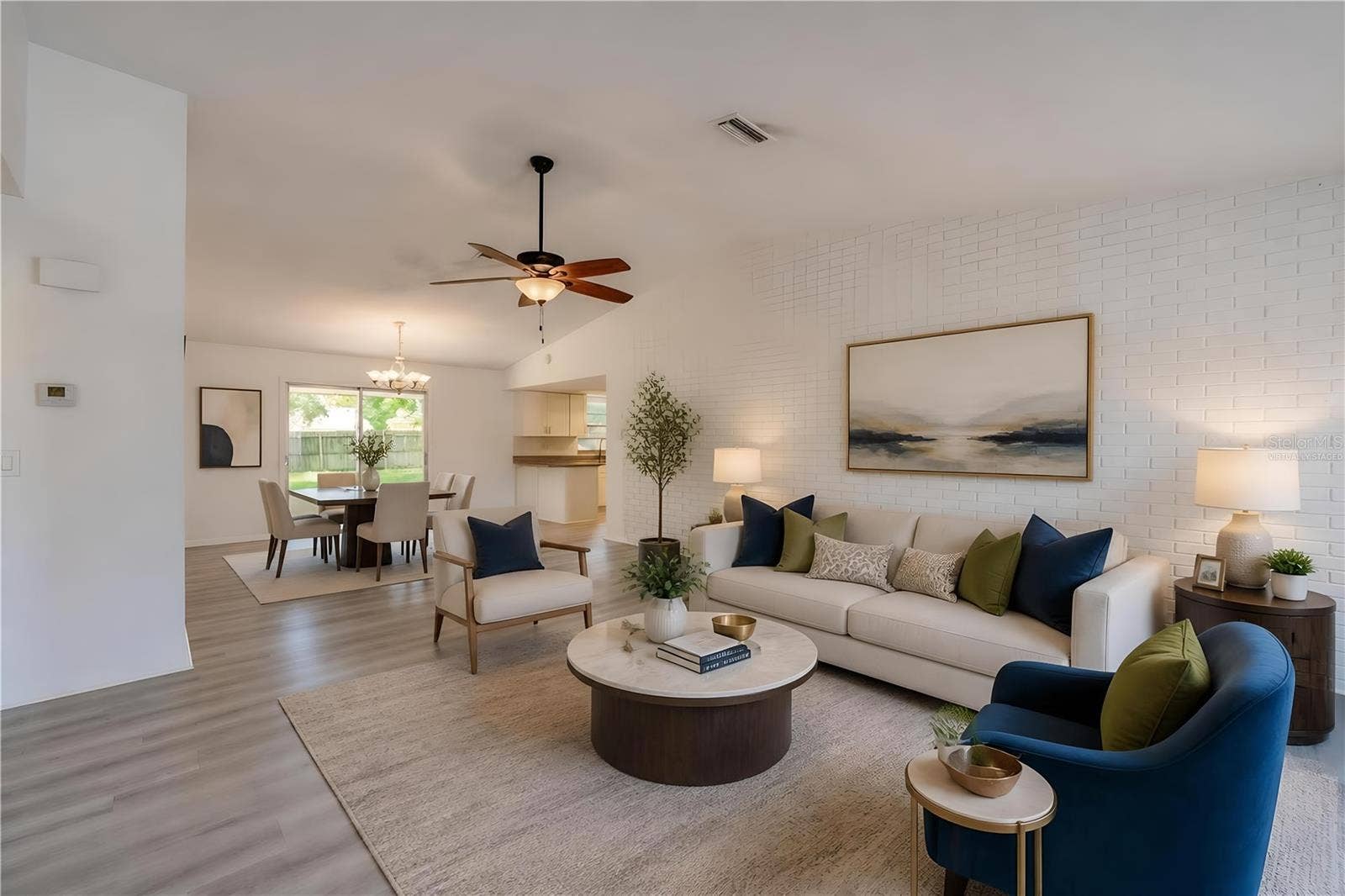 156 SPRINGWOOD DRIVE, Daytona Beach, FL 32119 Virtually Staged. Spacious open-concept living area highlighting light luxury vinyl flooring, a painted white brick accent wall, and a ceiling fan. Sliding glass doors at the rear provide seamless access and views to the backyard patio.