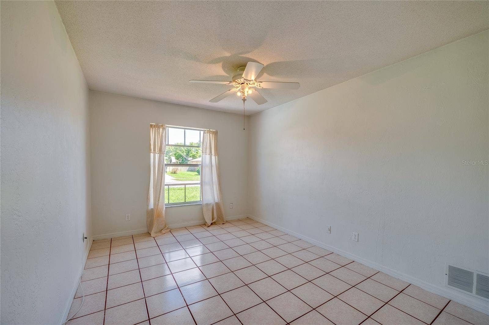 156 SPRINGWOOD DRIVE, Daytona Beach, FL 32119 Bright bedroom featuring light-colored tile flooring, neutral walls, and a ceiling fan with light fixture. A single window with curtains lets in natural light while providing views of the outdoors.