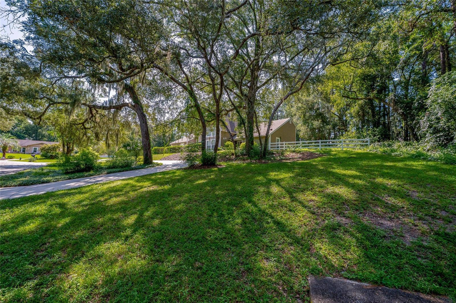 Expansive front lawn set beneath mature shade trees with a paved driveway and white fencing along the property line.