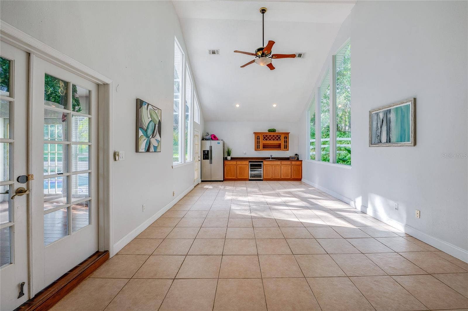 Spacious sunlit flex room featuring vaulted ceilings, a ceiling fan, wall-to-wall tile flooring, and floor-to-ceiling windows overlooking lush greenery.