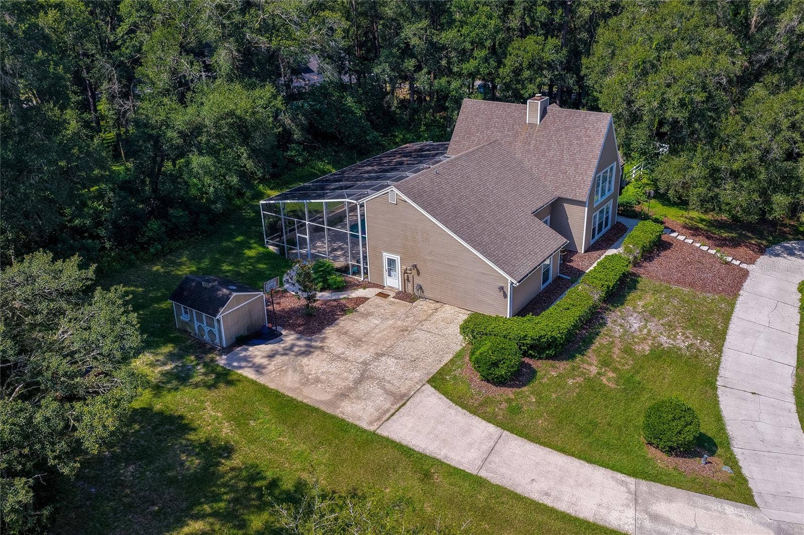 High-angle aerial view of the home showing its screened pool enclosure, side concrete parking pad, storage shed, and surrounding forest canopy.