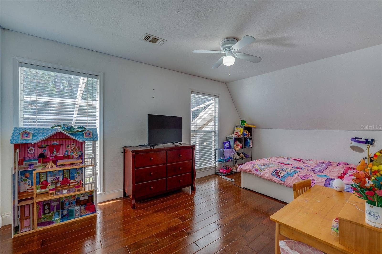 Bright bedroom featuring rich laminate flooring, a central ceiling fan, and sloped ceiling accents with dual windows.