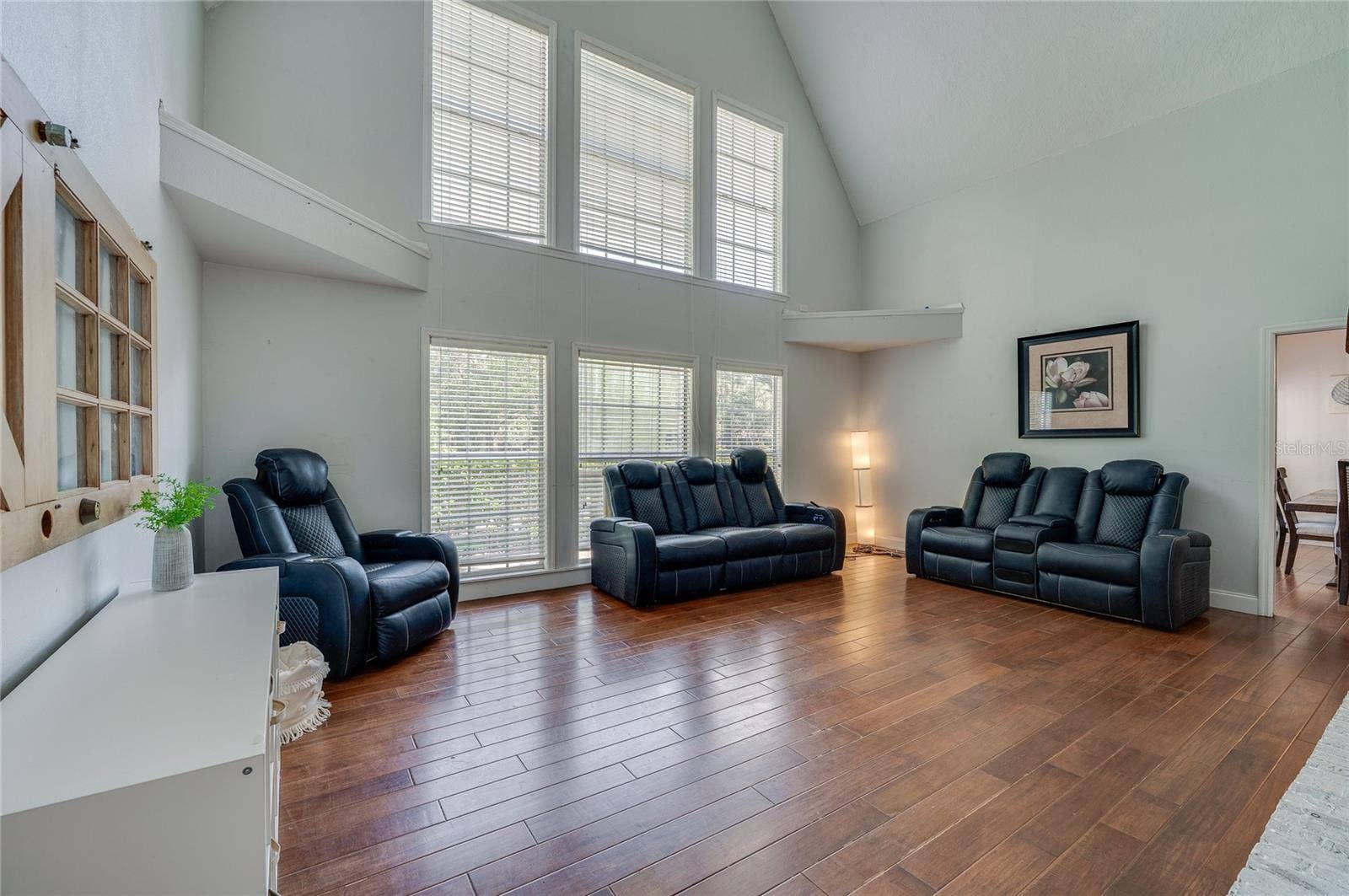 Grand living room highlighted by soaring vaulted ceilings, warm laminate floors, and a double-stacked bank of large windows offering abundant natural light.