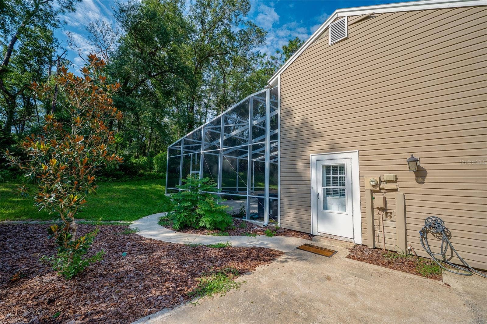 Rear exterior view of the home featuring tan vinyl siding, a paved walkway, landscaped mulch beds, and an attached screened-in enclosure.