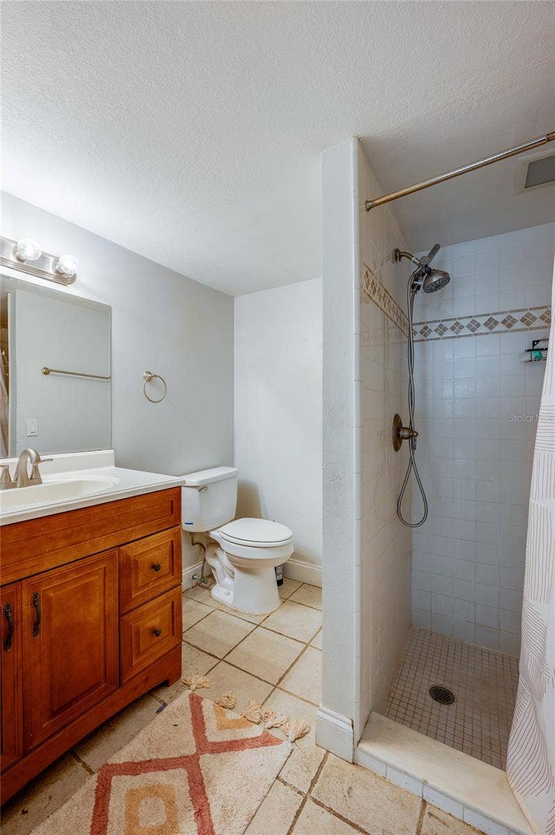 Compact bathroom with a tiled walk-in shower, warm wood vanity, single sink, and neutral ceramic tile flooring.