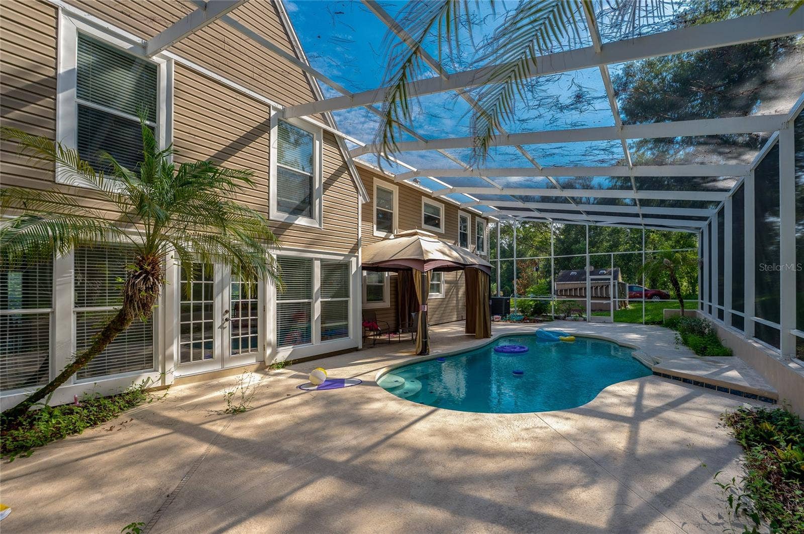 Sunlit outdoor pool deck enclosed in a screened lanai, highlighting French doors leading inside and lush tropical landscaping.