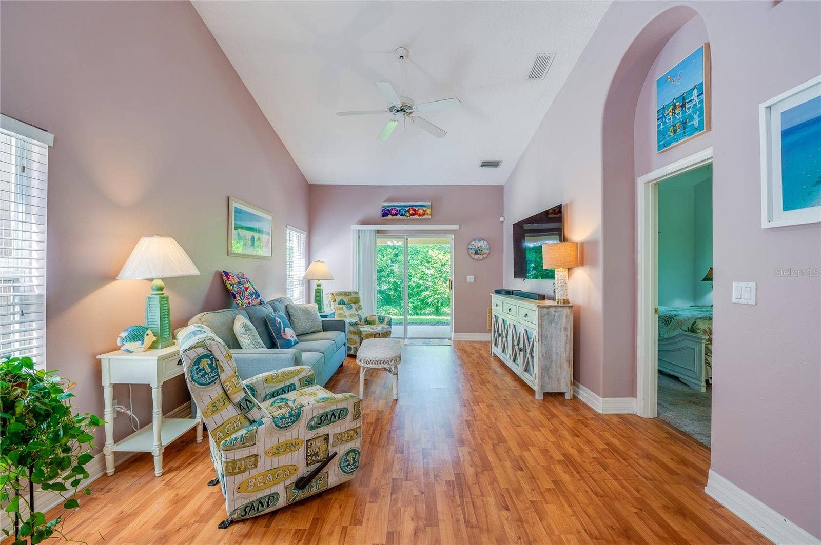 Spacious living room boasting dramatic vaulted ceilings, a central ceiling fan, and warm laminate flooring. Large sliding glass doors open directly to the outdoor patio space, while an arched doorway provides access to an adjoining bedroom.