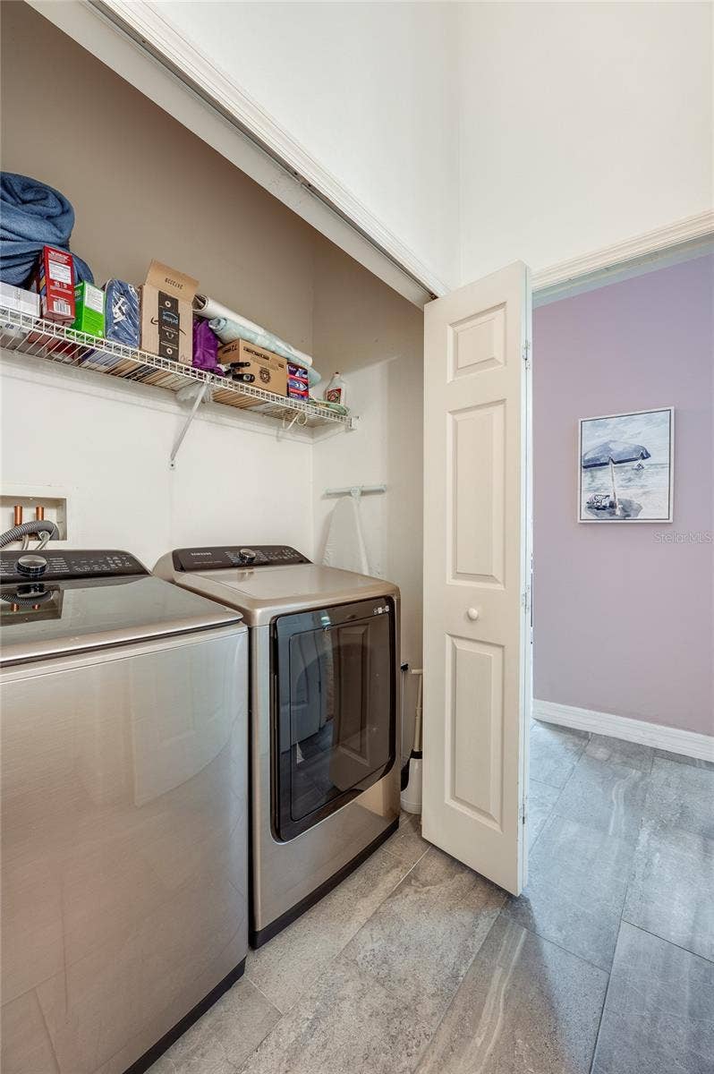 Dedicated laundry alcove equipped with side-by-side stainless steel appliances and overhead wire storage shelving. Grey tile flooring connects the utility space to the adjacent living areas.
