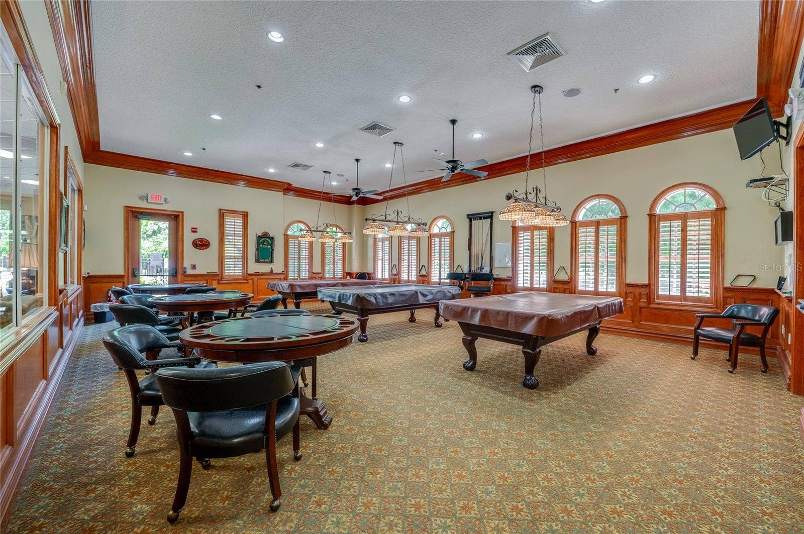 Spacious game and billiards room featuring recessed lighting, warm wood crown molding, and wainscoting throughout. Tall arched windows with wooden shutters illuminate the room, which connects to adjacent common spaces via large interior viewing windows.