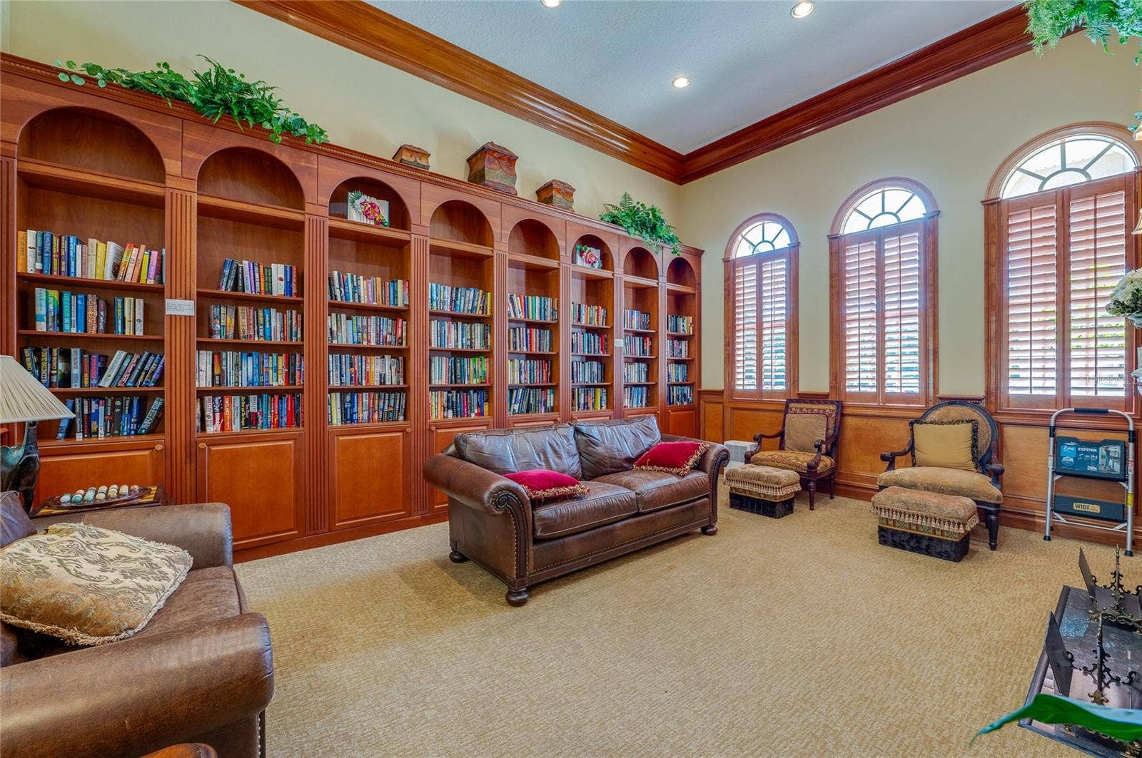 Cozy reading library framed by floor-to-ceiling rich wood built-in bookcases, warm crown molding, and neutral carpeting. Three large arched windows outfitted with wooden plantation shutters bring soft natural light into the space.