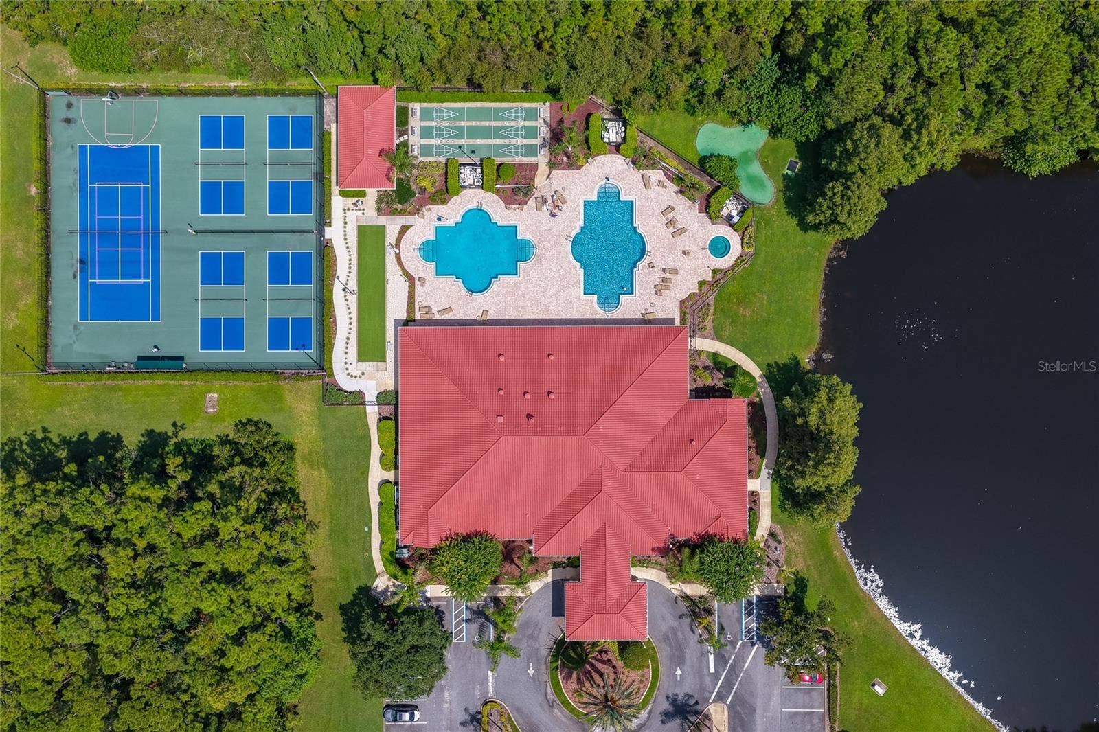 Top-down aerial view of community amenities including a spacious clubhouse with a distinct red roof, dual swimming pools, shuffleboard courts, and tennis courts. The facility is framed by a serene body of water and dense natural greenery.