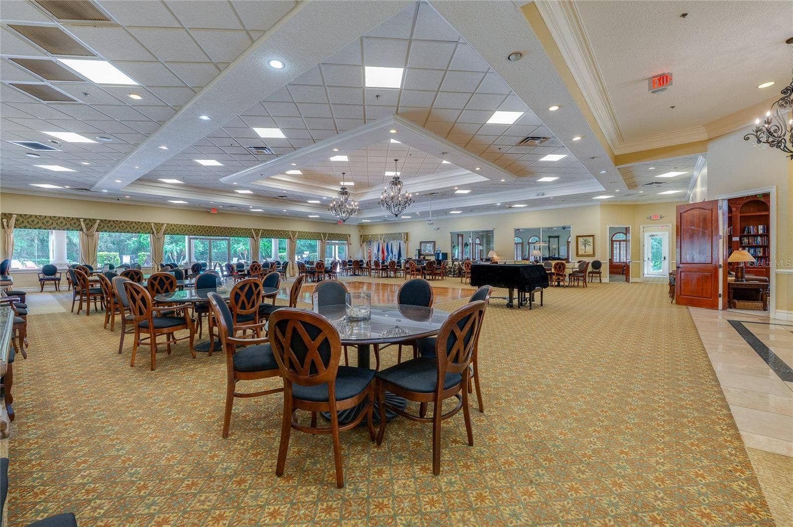 Expansive multi-purpose ballroom showcasing continuous patterned carpeting, multi-tiered tray ceilings with chandeliers, and a central polished dance floor. An open doorway on the side provides direct access to a quiet adjoining library space.