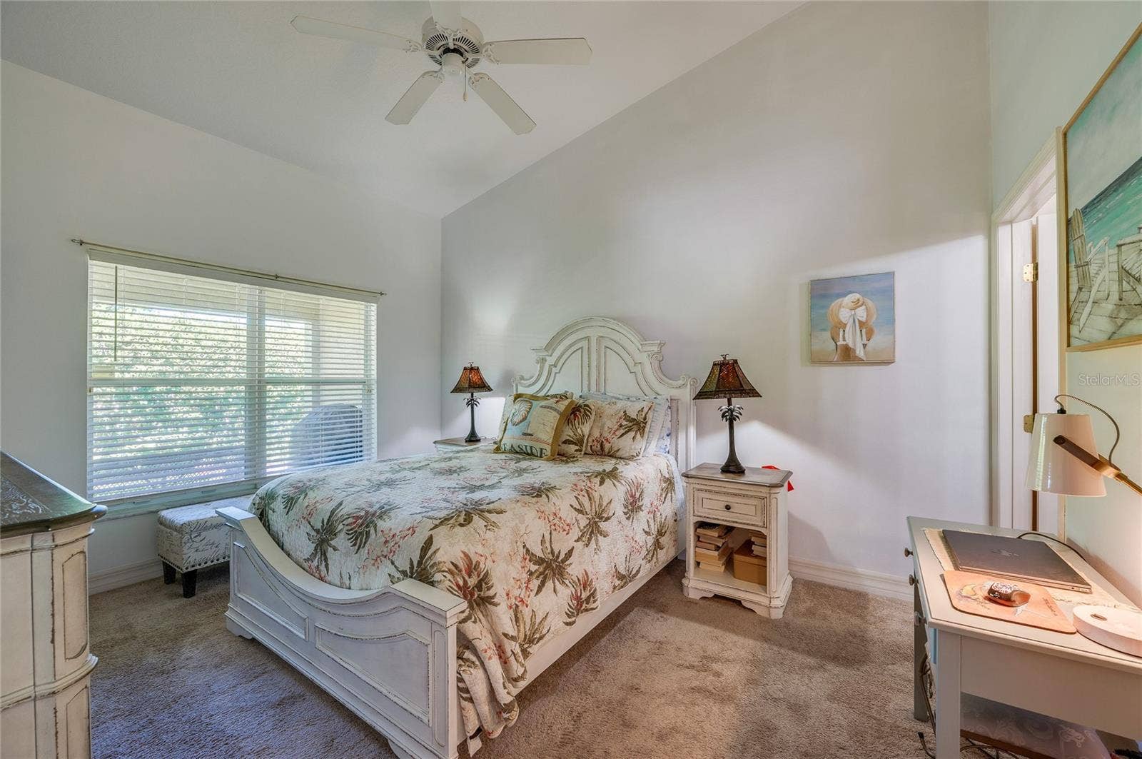 Serene bedroom suite featuring vaulted ceilings, soft neutral wall tones, and plush carpeting. A large window with horizontal blinds fills the room with indirect light.