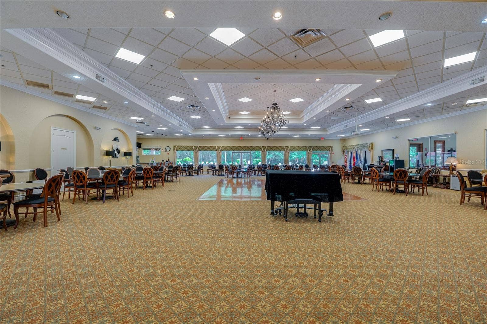 Grand event hall and dining room featuring drop tray ceilings, ornate chandelier lighting, patterned carpeting, and arched wall niches. Large glass doors along the far wall look out toward lush green landscaping.