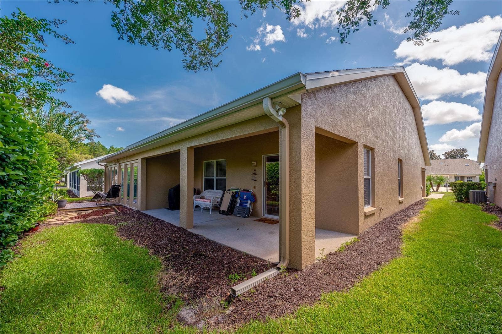 Exterior view of the rear elevation featuring a long covered patio and a well-maintained lawn bordered by mulch beds. Crisp stucco finishes and integrated roof gutters complete the exterior structure under clear blue skies.