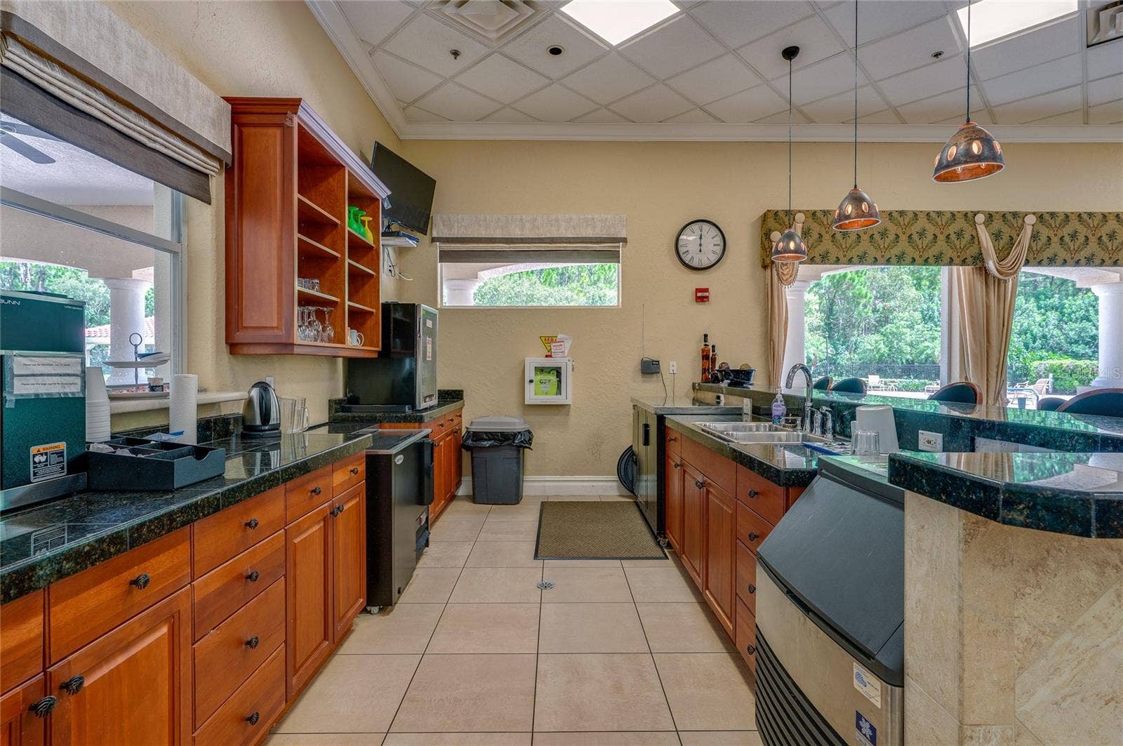 Functional beverage station and service kitchen equipped with extensive dark granite countertops, rich wood cabinetry, stainless steel sink stations, and tile flooring. Wide windows and valanced drapery overlook the outdoor grounds.