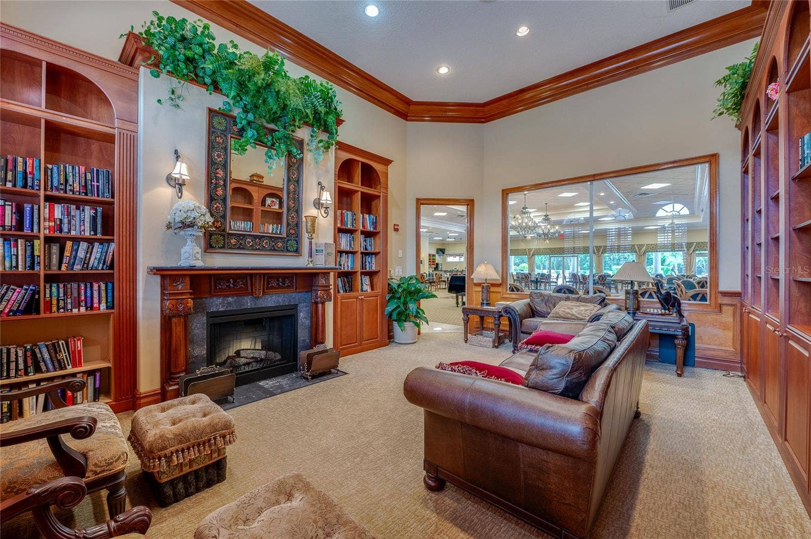 Warmly styled library corner highlighted by a classic wood-mantel fireplace with a stone surround, vibrant wall greenery, and custom built-in shelving. A large interior window cut-out looks directly through into the main event hall.