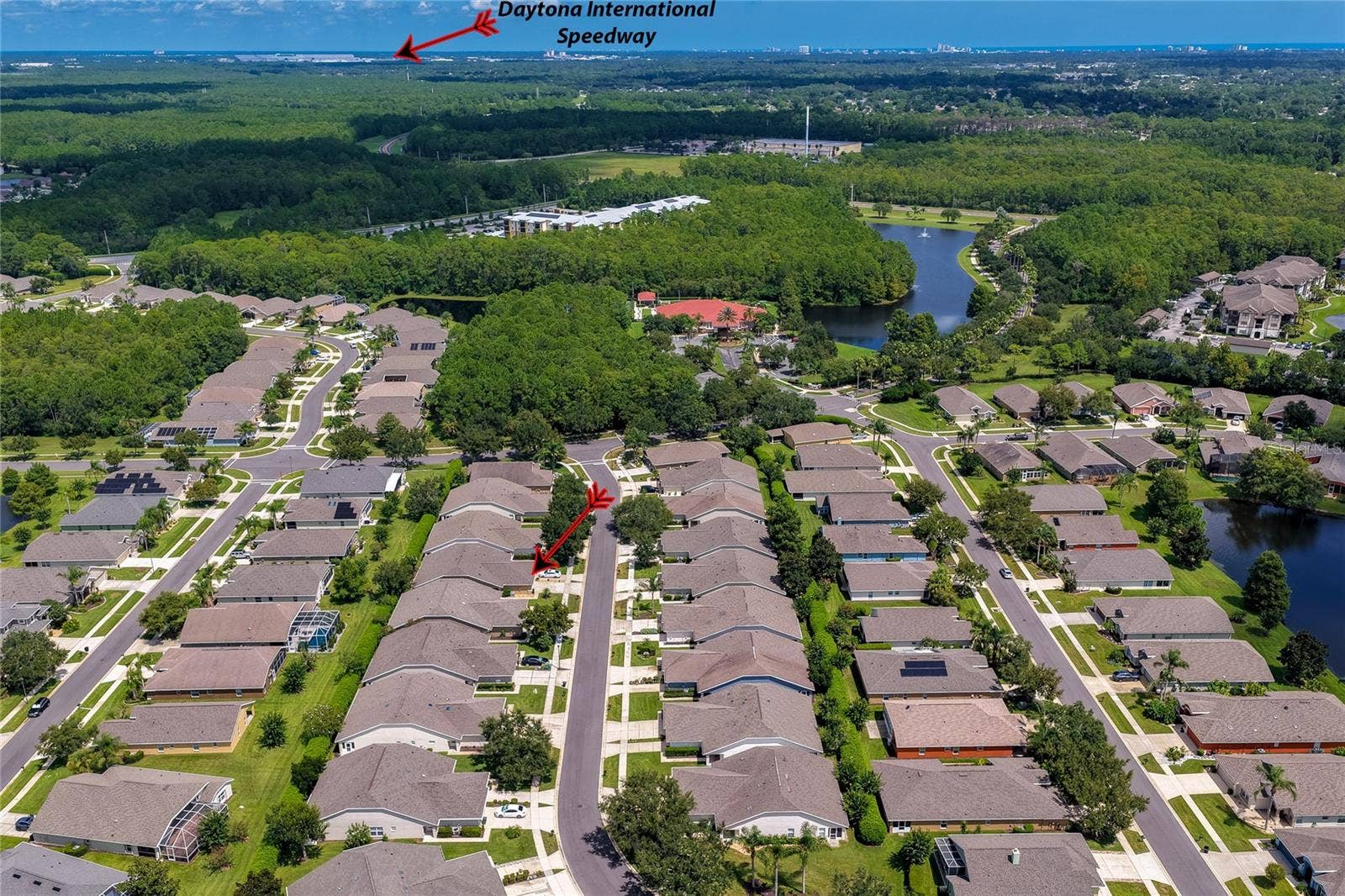 High-altitude aerial view showcasing the expansive residential development surrounded by dense woodlands, with a directional arrow pointing toward the iconic Daytona International Speedway in the distance.