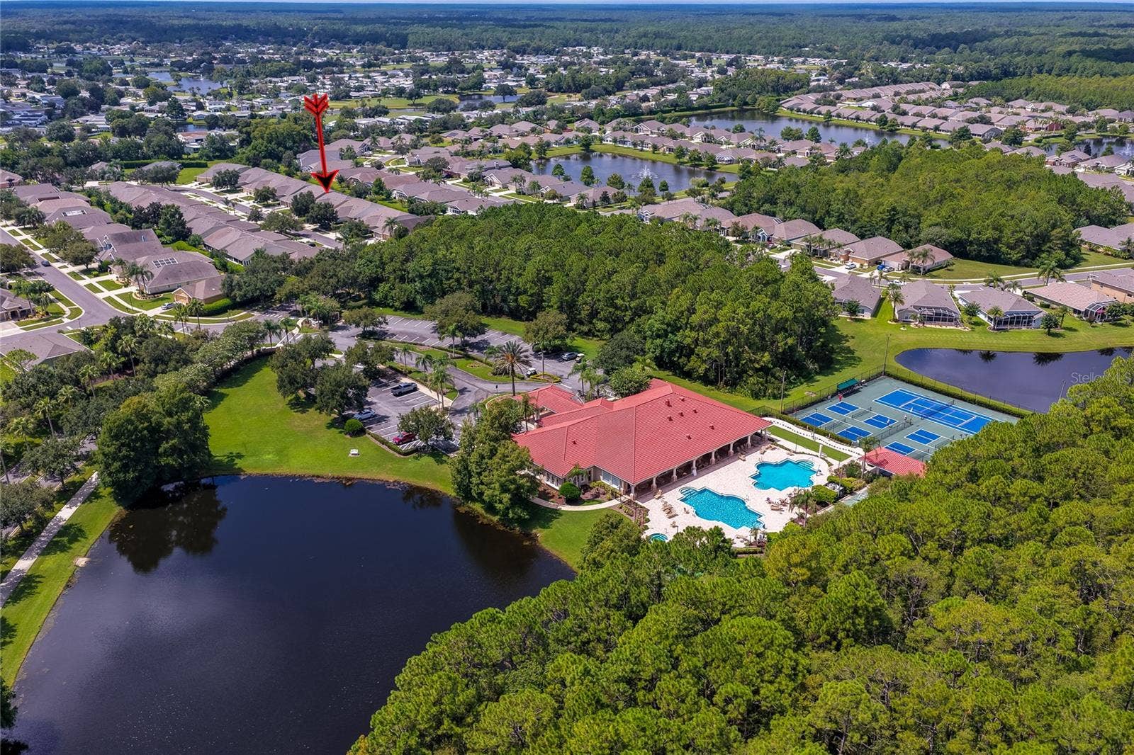 Elevated wide-angle aerial perspective highlighting the community clubhouse, sports courts, and adjacent lake, with an arrow pointing toward the neighborhood homes nestled behind green tree lines.