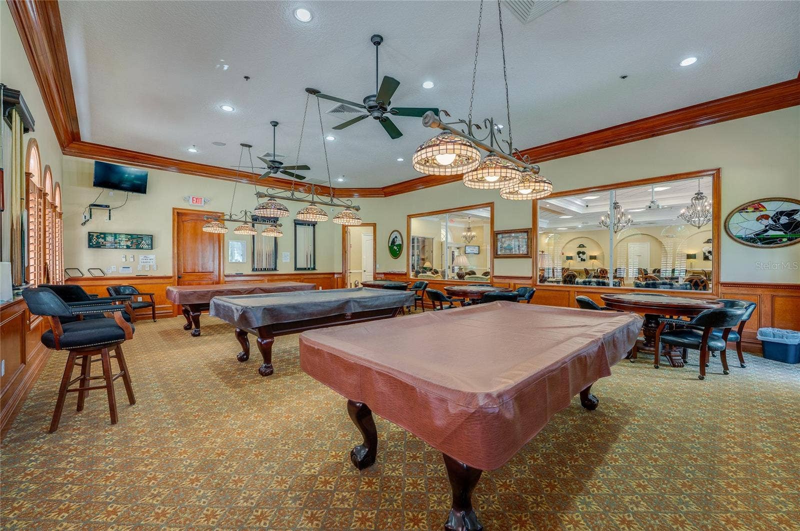 Secondary angle of the game room showcasing detailed wood wall trim, hanging stained-glass pendant lights, and overhead ceiling fans. An interior glass partition window provides a clear view into the main hall.