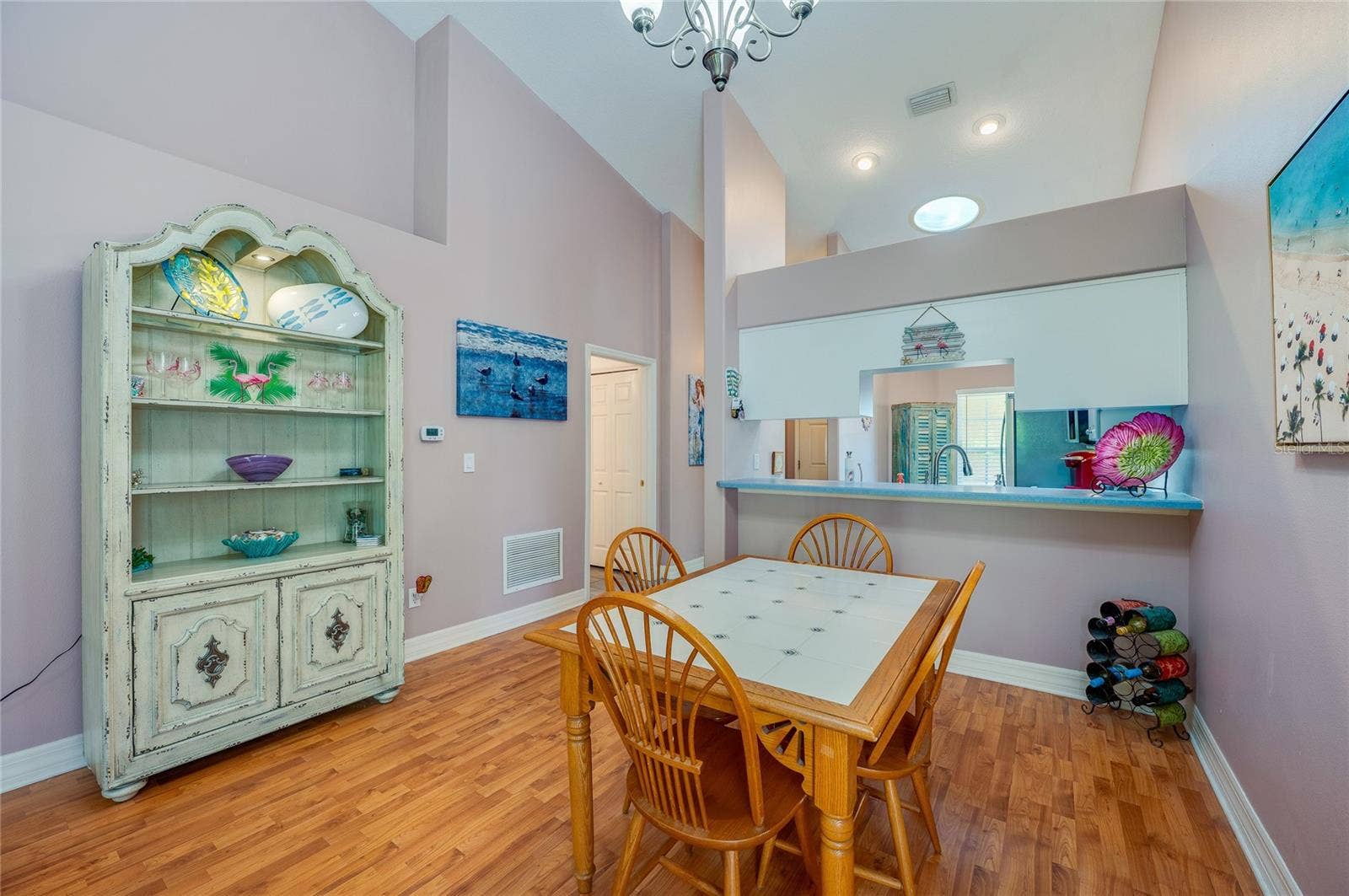 Light-filled dining space featuring high vaulted ceilings, modern overhead lighting, and rich laminate flooring. A low partition wall with a pass-through counter offers a seamless connection directly into the kitchen.