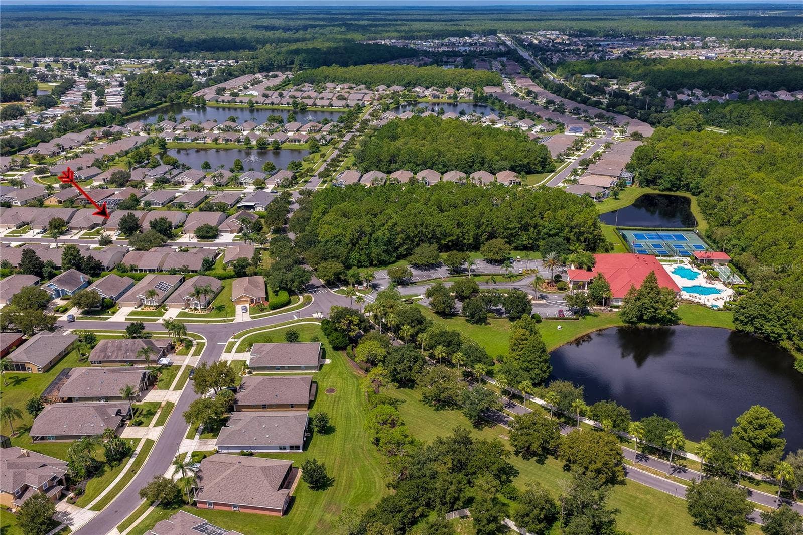 Sweeping neighborhood aerial displaying quiet residential streets lined with single-story homes, tranquil ponds, and lush green common areas. An indicator arrow marks a specific residence within the scenic layout.