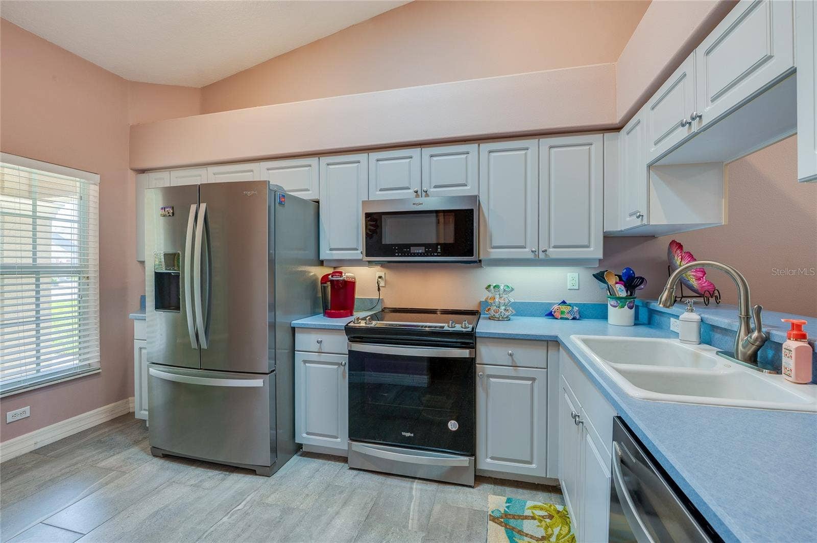 Functional kitchen equipped with stainless steel appliances, white cabinetry, and blue countertops. A large window brings in natural light, while high vaulted ceilings add depth.