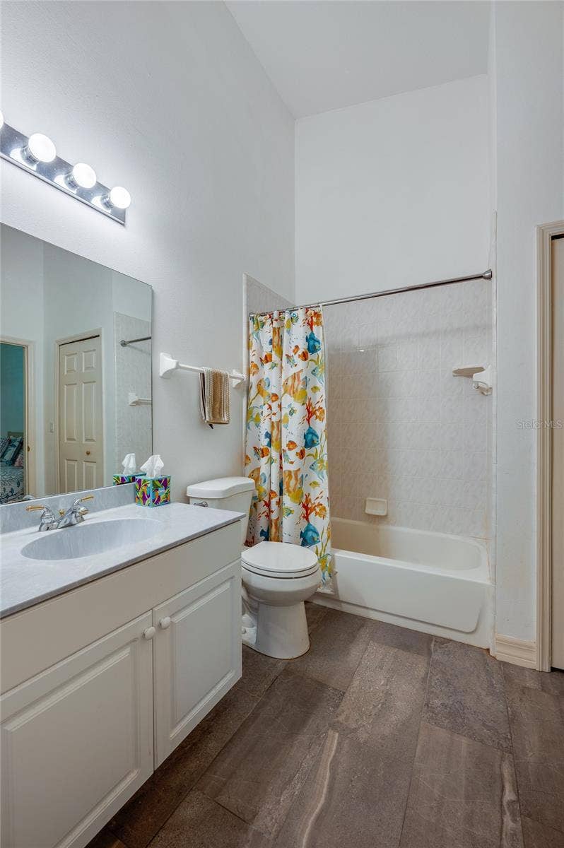 Functional bathroom featuring dark tile flooring, a wide vanity mirror, and bright overhead lighting. The combination shower and bathtub includes built-in wall shelving and tiled surrounds.