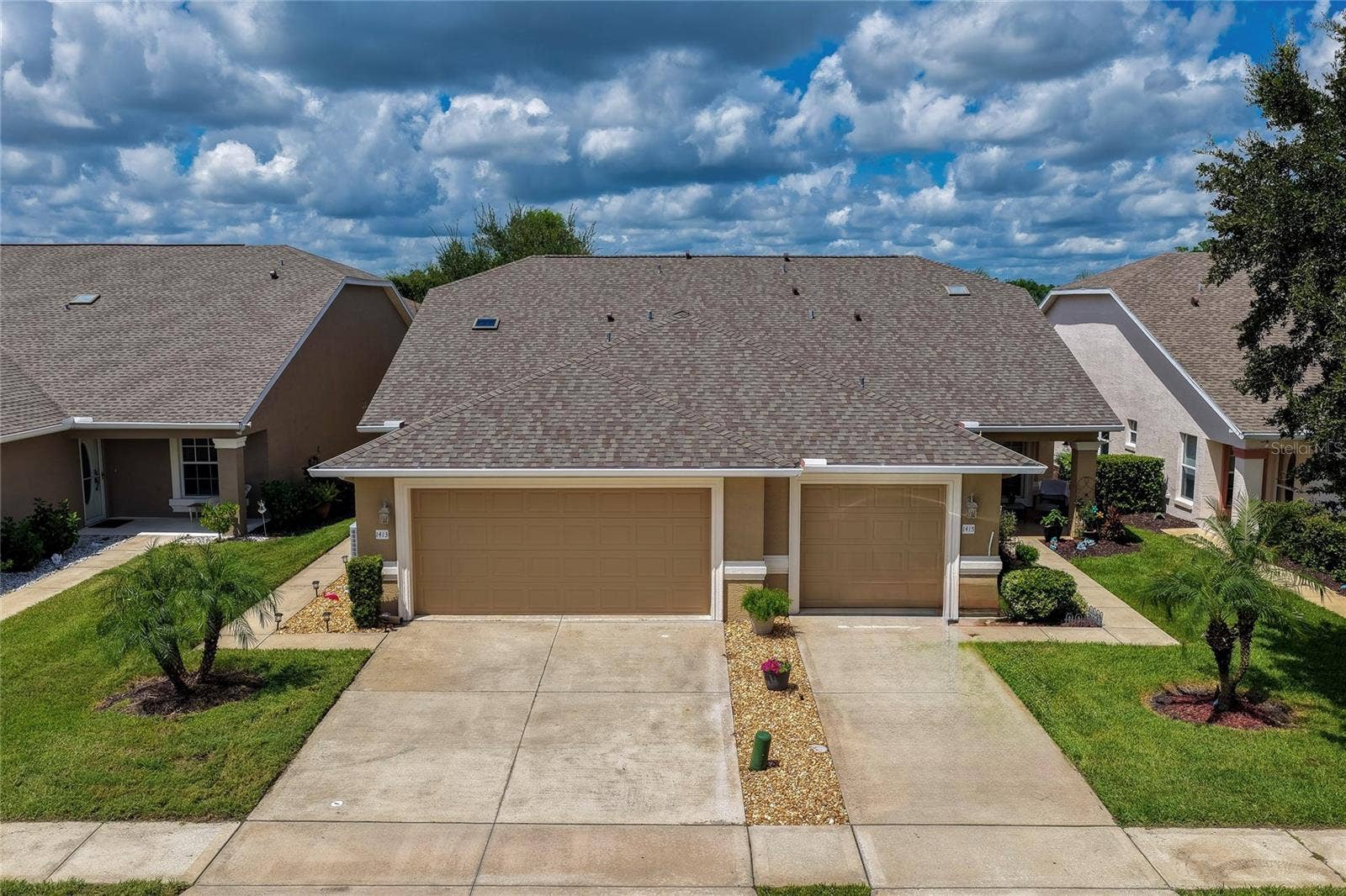 Front-facing view of the home featuring neutral stucco siding, an attached garage, and an expansive paved driveway surrounded by tropical green landscaping.