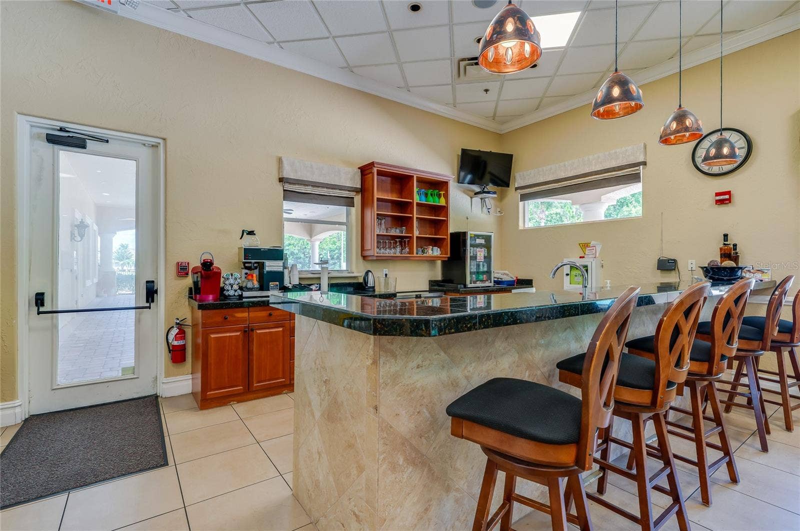 Clean service bar area featuring dark granite counter surfaces, warm wood cabinetry, neutral tile flooring, and industrial-style hanging pendant lights. An exterior door opens directly to a paved covered walkway outside.