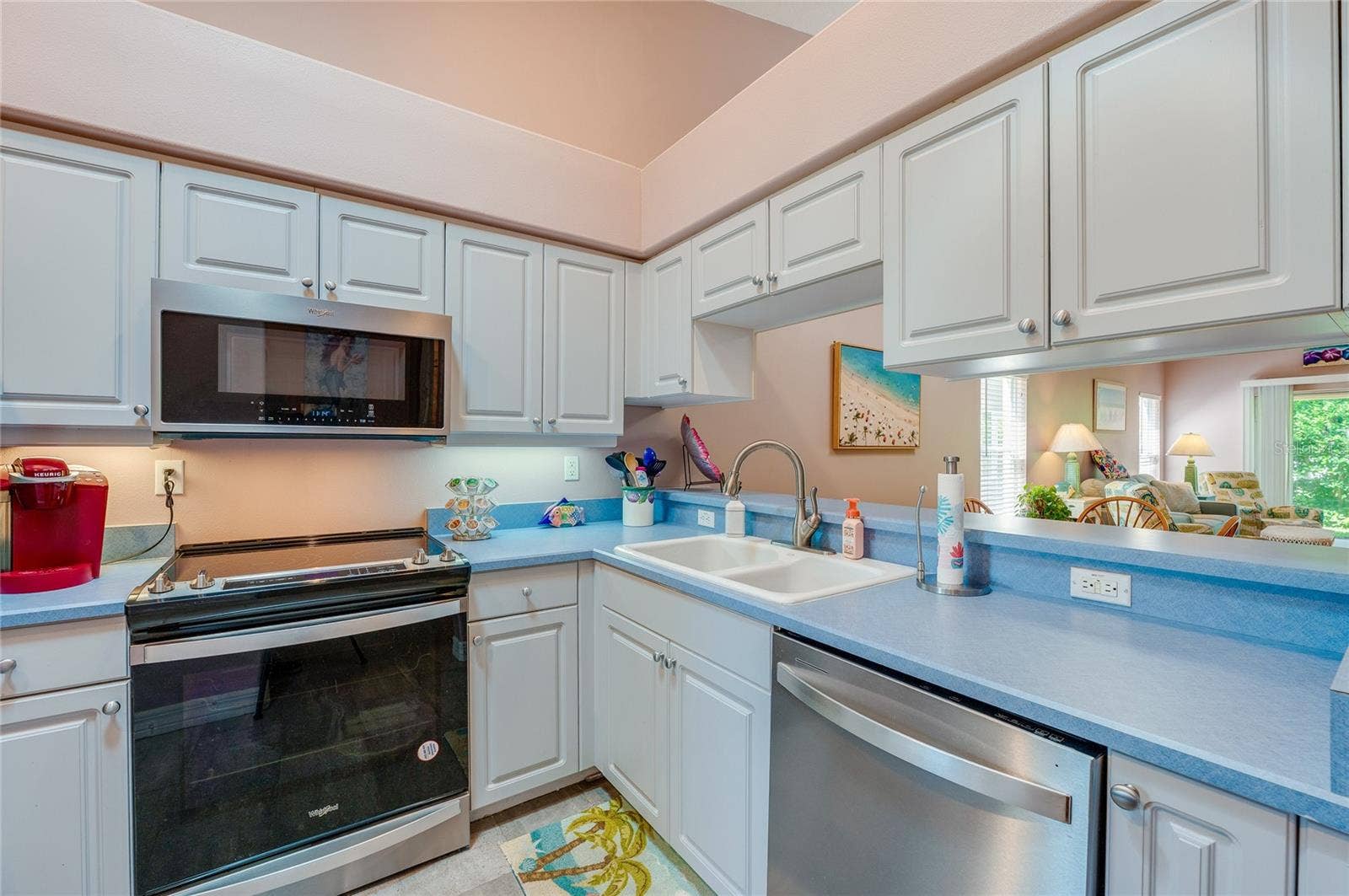 Efficient kitchen layout highlighting a built-in dishwasher, electric range, microwave, and double-basin sink. A pass-through window maintains a seamless connection to the adjacent living area.