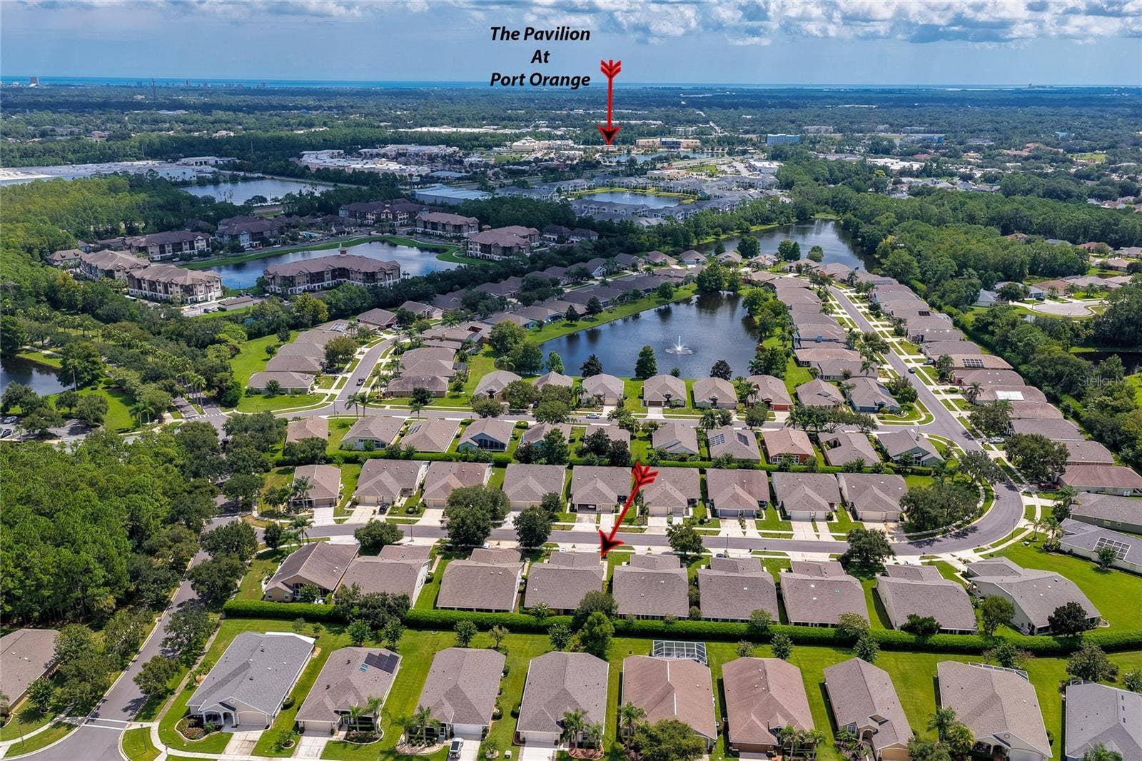 Broad aerial view capturing the residential neighborhood layout, surrounding water features, and green foliage, with a red arrow pointing toward the nearby Pavilion at Port Orange shopping area.