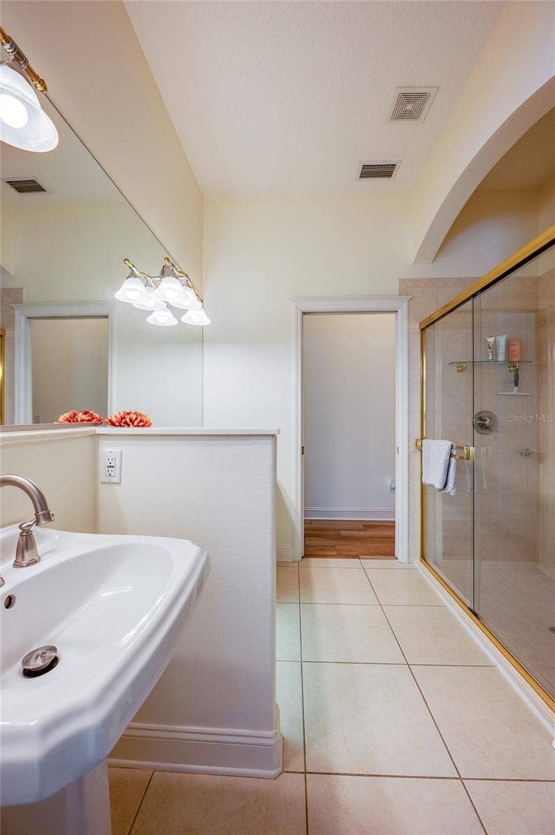 25 REMINGTON ROAD, Ormond Beach, FL 32174 Clean bathroom space featuring a white pedestal sink, a glass-enclosed walk-in shower with gold trim, and light tiled flooring throughout.