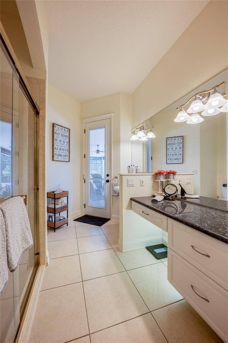 25 REMINGTON ROAD, Ormond Beach, FL 32174 Bright bathroom featuring neutral floor tile, a glass-enclosed shower, and a expansive mirror above the vanity with dark countertops. A door provides direct access to the outdoors.