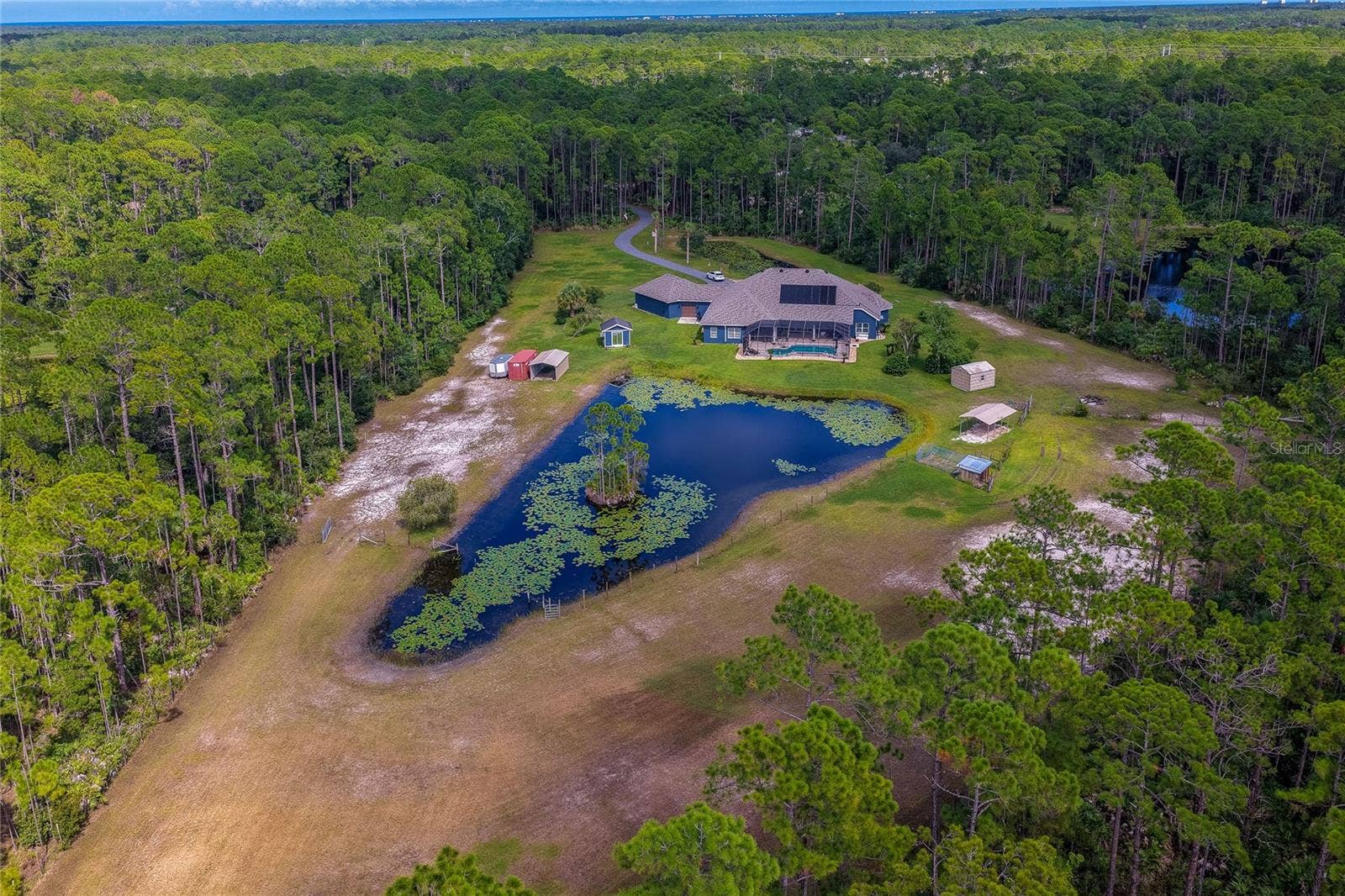 25 REMINGTON ROAD, Ormond Beach, FL 32174 High-altitude aerial shot displaying the private estate complete with its custom home, enclosed pool, large lily pad pond, outbuildings, and dense forest surroundings.