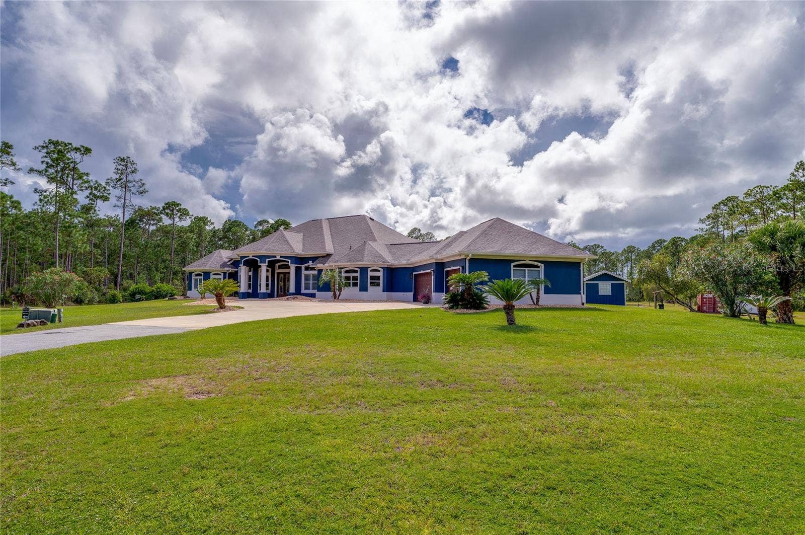 25 REMINGTON ROAD, Ormond Beach, FL 32174 Spacious front view of a custom blue home featuring architectural white columns, an expansive driveway, and neatly landscaped palm trees set against a backdrop of mature woods.