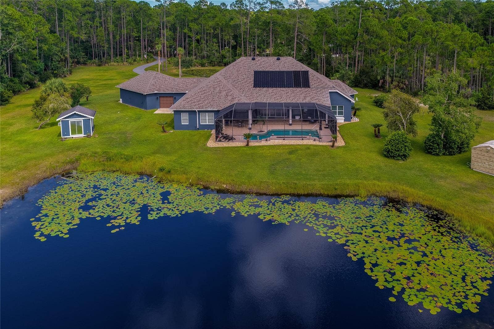 25 REMINGTON ROAD, Ormond Beach, FL 32174 Elevated rear view of the home highlighting the screened pool enclosure, roof-mounted solar panels, sheds, and direct access to the private lily pad pond.