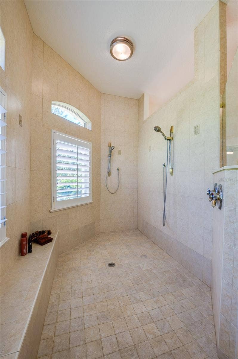 25 REMINGTON ROAD, Ormond Beach, FL 32174 Large walk-in tile shower equipped with multiple shower heads, a built-in bench, and shuttered windows for natural light.