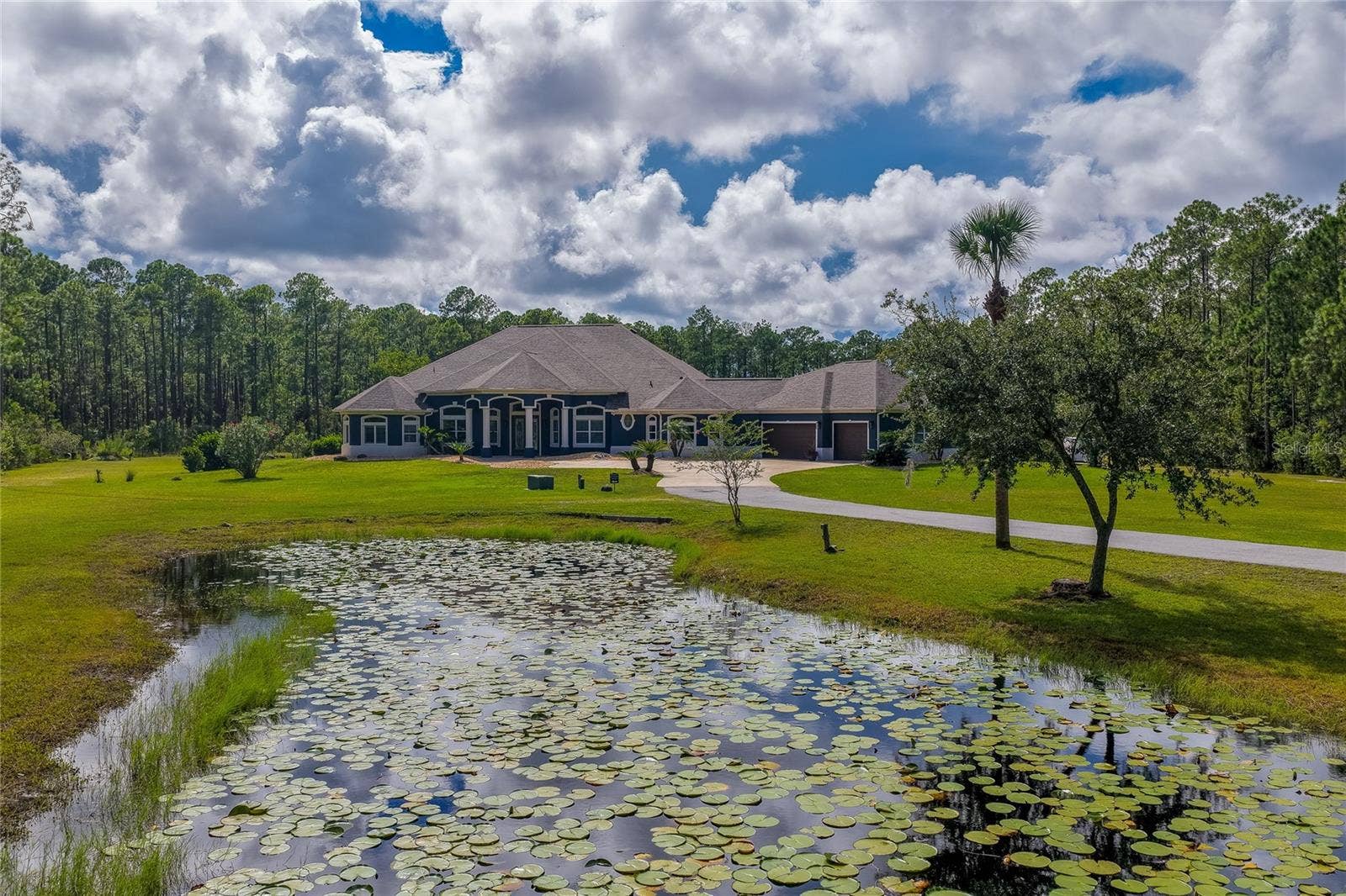 25 REMINGTON ROAD, Ormond Beach, FL 32174 Foreground view of a lily-pad-covered pond looking toward a sprawling home framed by a paved driveway and mature pine trees.