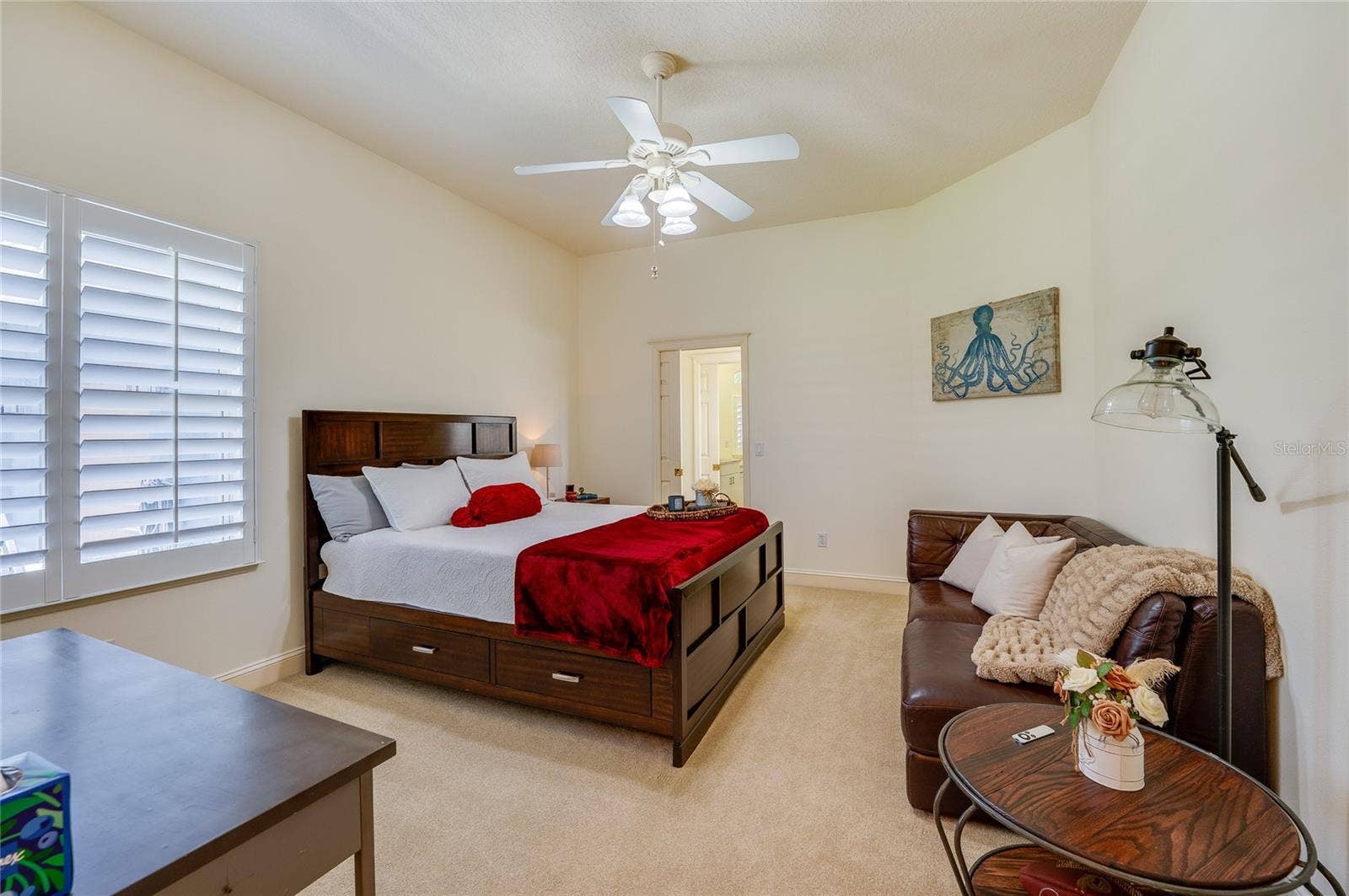 25 REMINGTON ROAD, Ormond Beach, FL 32174 Inviting bedroom layout featuring large windows fitted with plantation shutters, plush carpeting, and an ensuite doorway. A ceiling fan offers added ventilation.