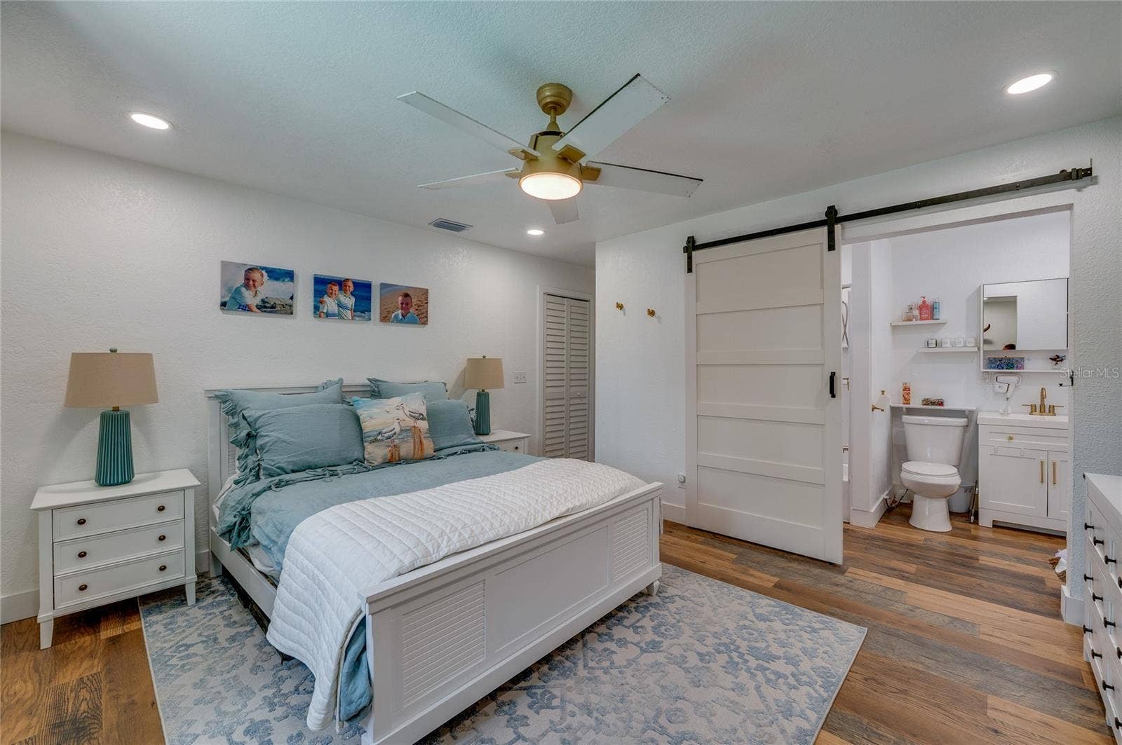 348 WOODLAND AVENUE, Daytona Beach, FL 32118 Spacious primary bedroom suite showcasing warm LVP flooring, a ceiling fan, and direct access to an ensuite bathroom through a sliding white barn door.