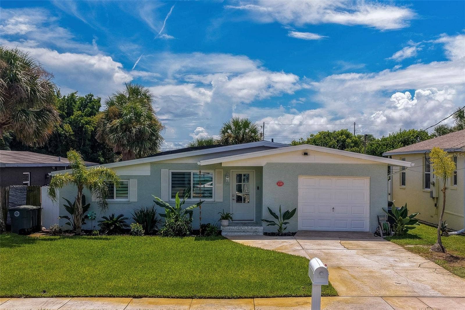 348 WOODLAND AVENUE, Daytona Beach, FL 32118 Charming single-story exterior showcasing a bright lawn, lush tropical landscaping, and a clean driveway leading to an attached garage under a vibrant blue sky.