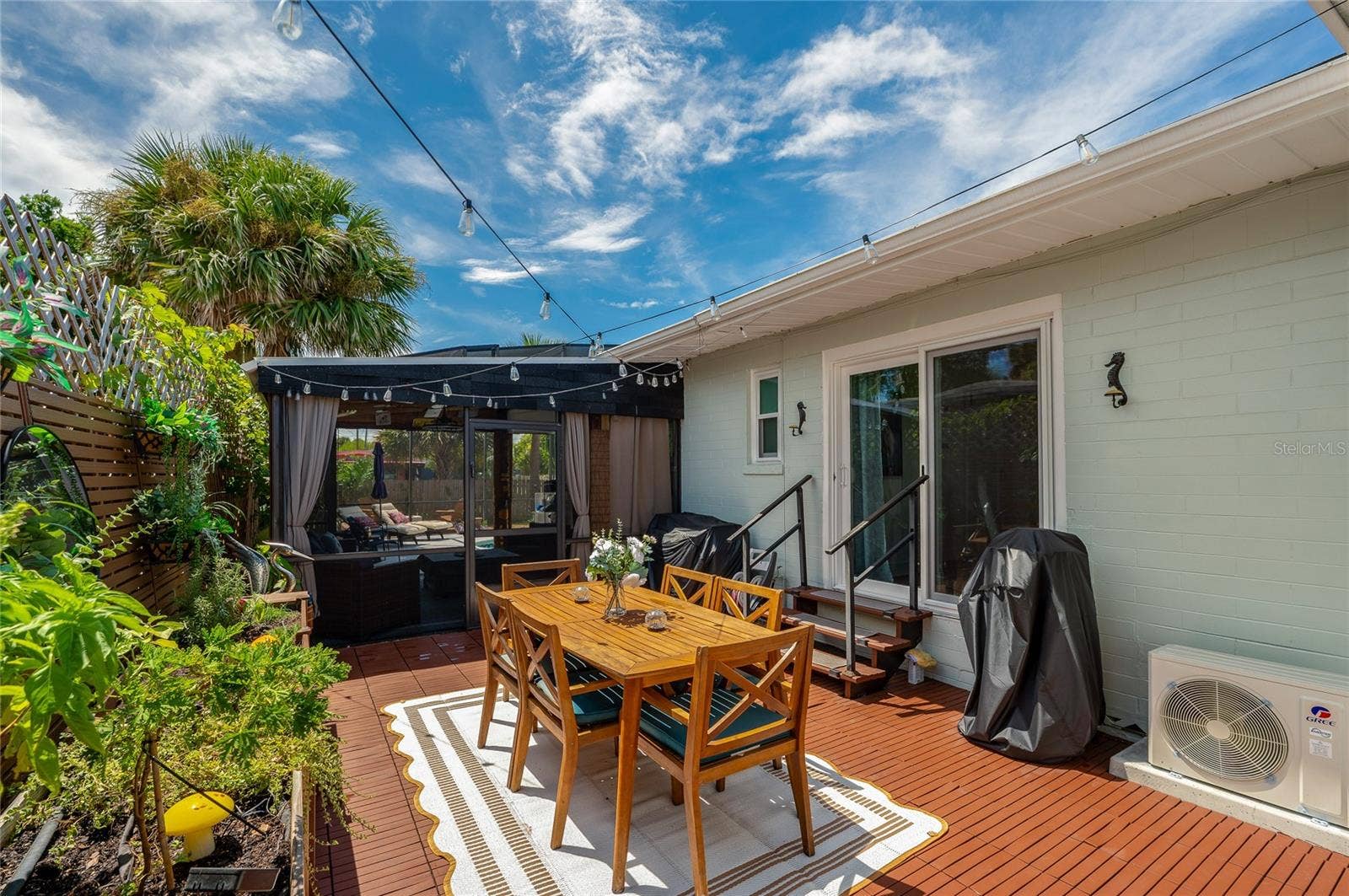 348 WOODLAND AVENUE, Daytona Beach, FL 32118 Spacious outdoor patio area with dark wooden decking, sliding glass door access, and string lights overhead. The space includes raised garden planters and a adjacent screen enclosure.