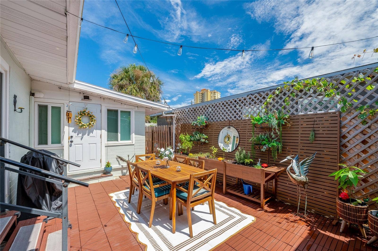 348 WOODLAND AVENUE, Daytona Beach, FL 32118 Charming outdoor wooden deck framed by lattice fencing, ambient string lights, and potted greenery. An entryway with a decorative wreath adds a welcoming touch.