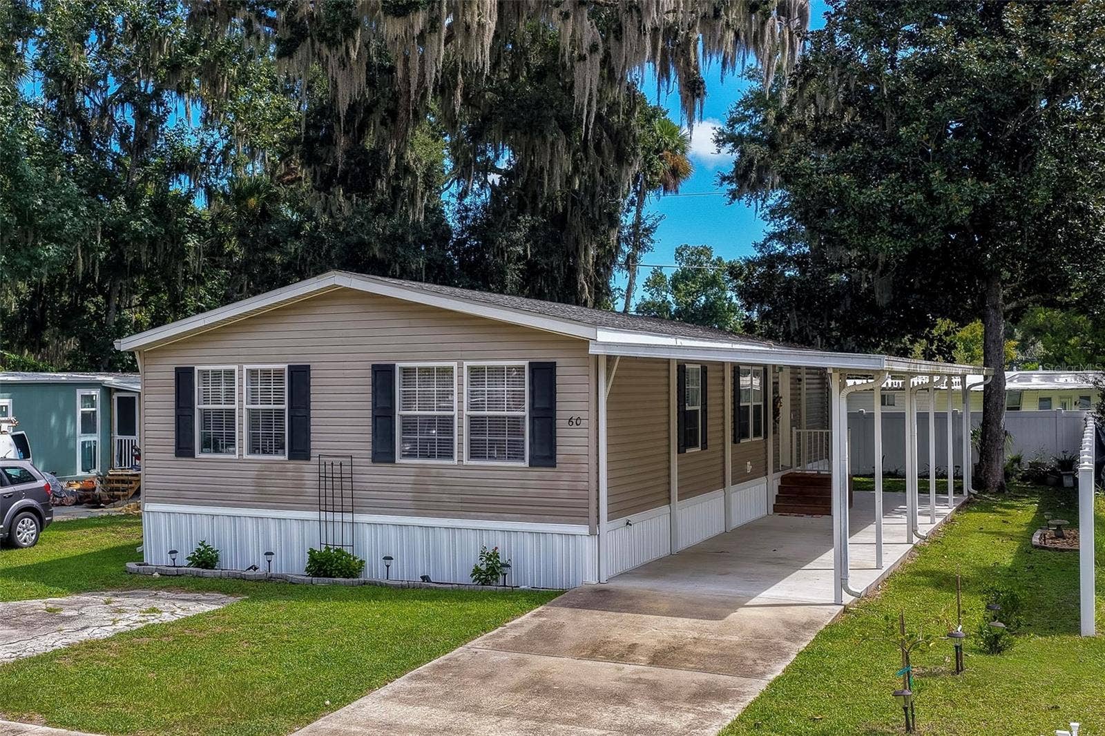 60 GOLDEN GATE CIRCLE, Port Orange, FL 32129 Charming exterior view of a single-story home featuring vinyl siding, dark shutters, and a long covered carport driveway. The property is framed by mature, Spanish moss-draped trees and neat lawn fencing.