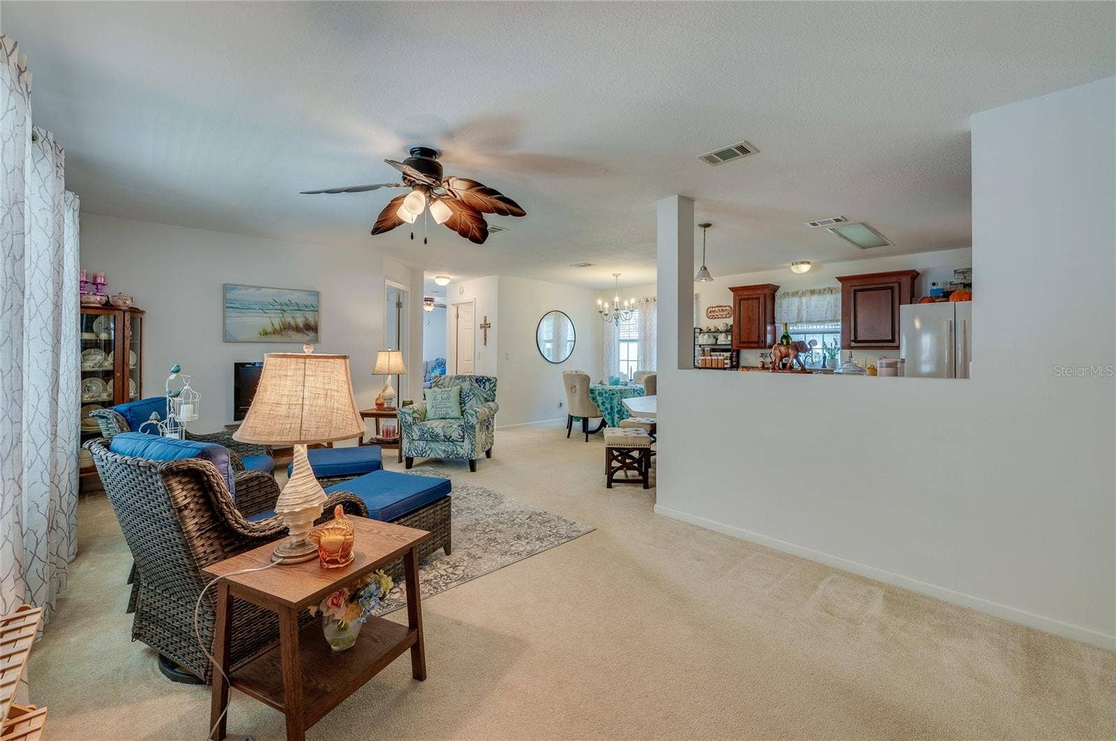 60 GOLDEN GATE CIRCLE, Port Orange, FL 32129 Bright and spacious open-concept living area featuring soft neutral wall tones, low-maintenance carpeting, and ceiling fan fixture. A half-wall seamlessly connects the main living space with the adjacent kitchen area.